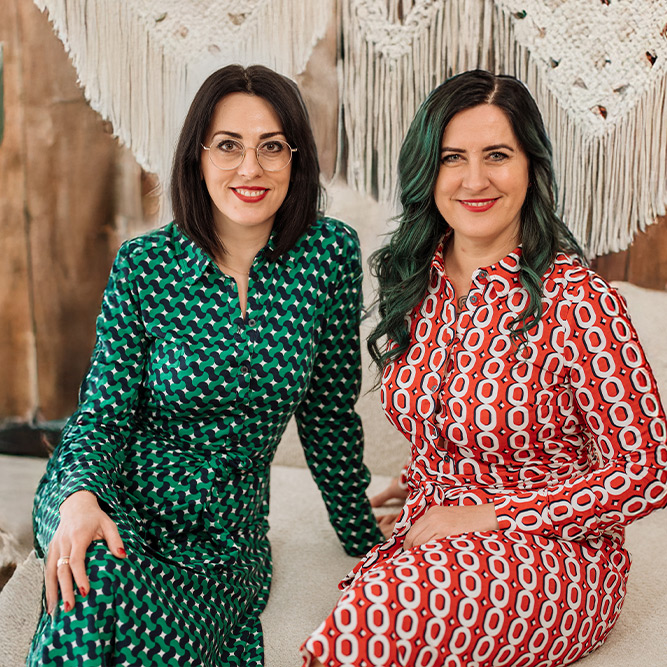 Olga Geipel links und Nelly Rühle rechts, sitzen in gemusterten Kleidern nebeneinander auf einem beigen Sofa mit einem Boho-Makamee Hintergrund